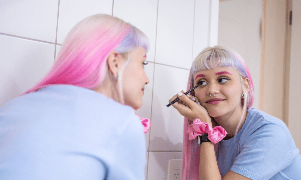 a young woman with pink hair applying make up in the mirror