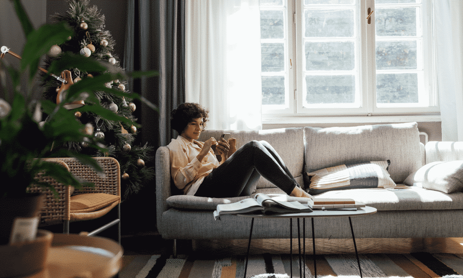 a woman sitting on a couch looking at her phone with a christmas tree in the corner